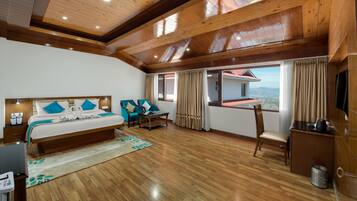 A hotel room with a wooden ceiling, a bed with blue pillows, a desk, a chair, and a view of a house through the window.