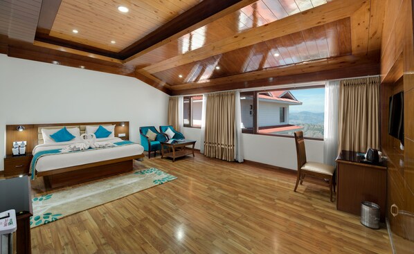 A hotel room with a wooden ceiling, a bed with blue pillows, a desk, a chair, and a view of a house through the window.