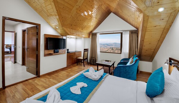 Habitación de hotel con techo de madera, cama con ropa de cama azul y blanca, televisor montado en la pared, escritorio con silla y vista a las montañas por la ventana.