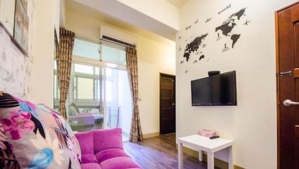 Una sala de estar con un sofá rosa, una mesita blanca pequeña, un televisor montado en la pared y una puerta que da a otra habitación.