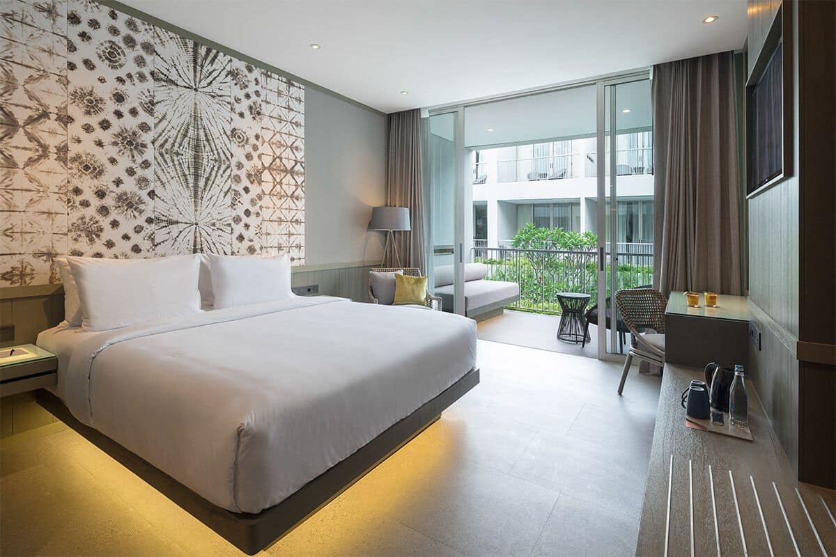 A modern hotel room with a large bed, a balcony with seating, and a view of the building's exterior.