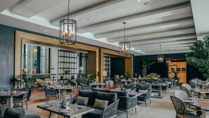 A spacious indoor dining area with multiple tables and chairs, large windows, and hanging pendant lights. - Kanvaz Village Resort Seminyak (Keroboken Kelod)
