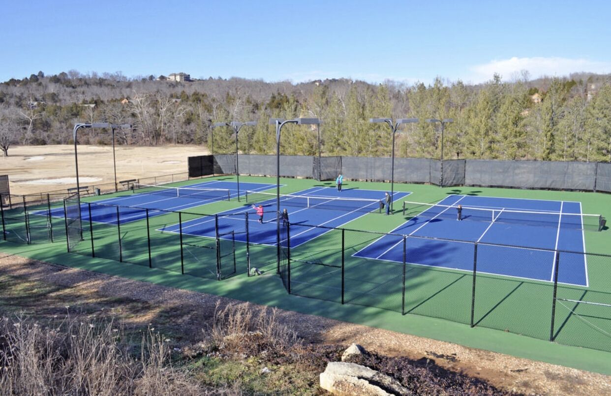 An outdoor tennis court complex with multiple courts, surrounded by a fence and trees.