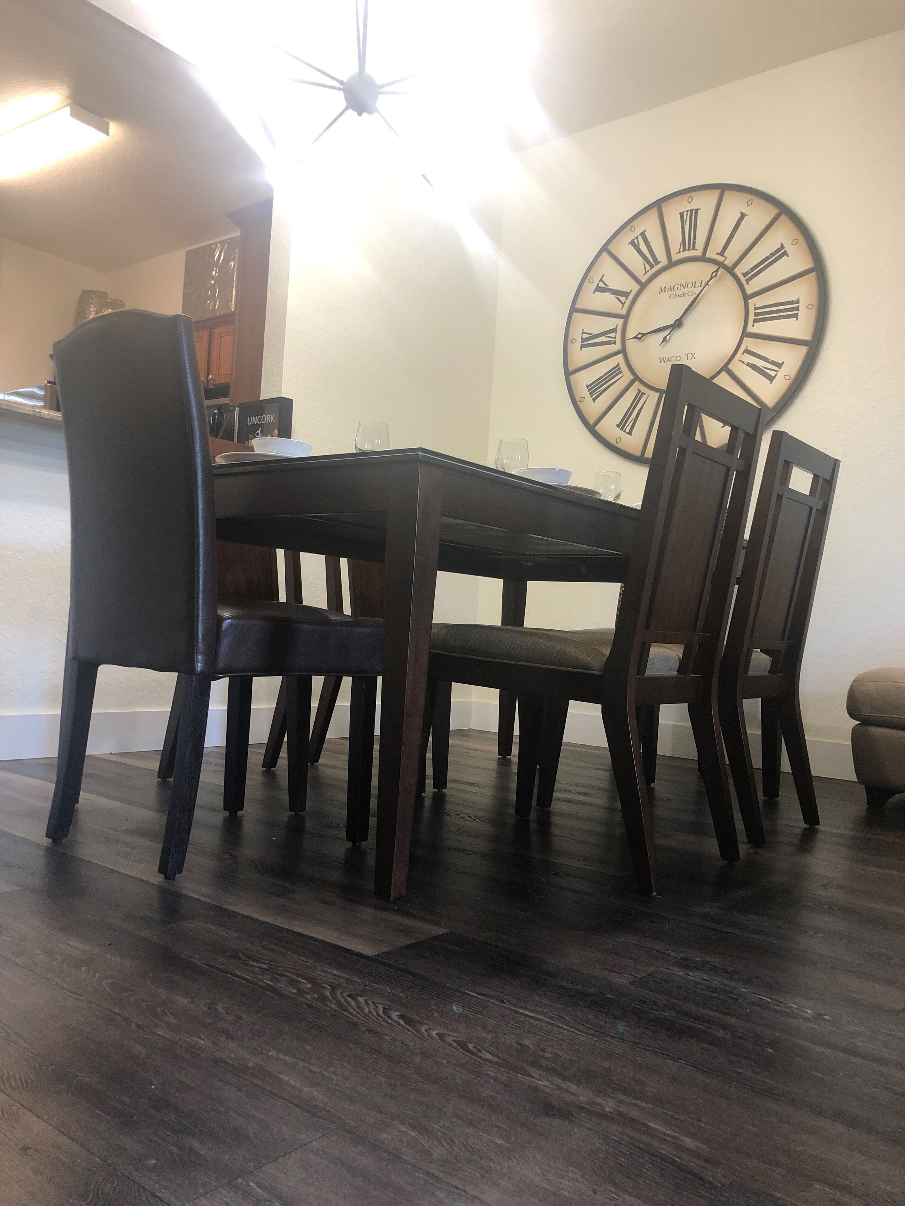 A dining area with a wooden table and chairs, a large wall clock, and a kitchen in the background.