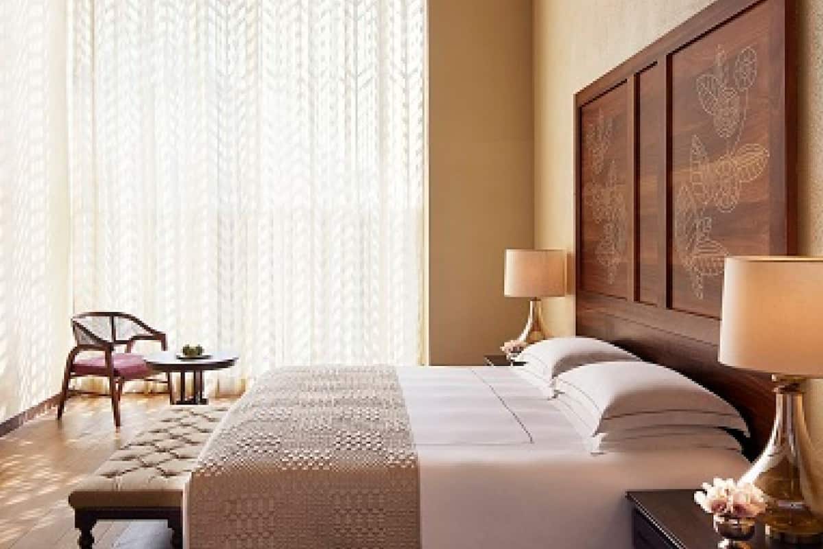 A hotel room with a large bed, two bedside lamps, a wooden headboard with floral carvings, a small table with a chair, and sheer curtains.
