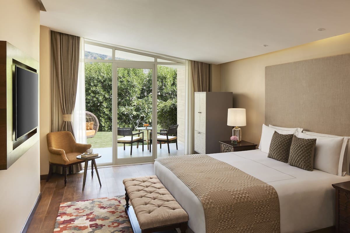 A hotel room with a large bed, a sitting area with a chair and table, a television, and a view of a garden through the sliding doors.