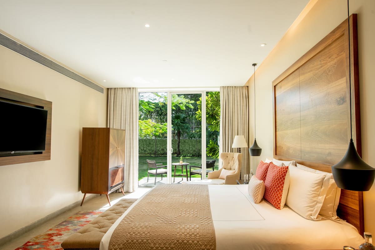 A modern hotel room with a large bed, a flat-screen TV, a wooden cabinet, and a view of a garden through a sliding glass door.