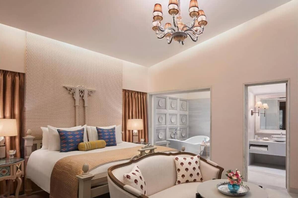 A hotel room with a bed, a sofa, a round table, a chandelier, a bathroom with a bathtub, and a large mirror.