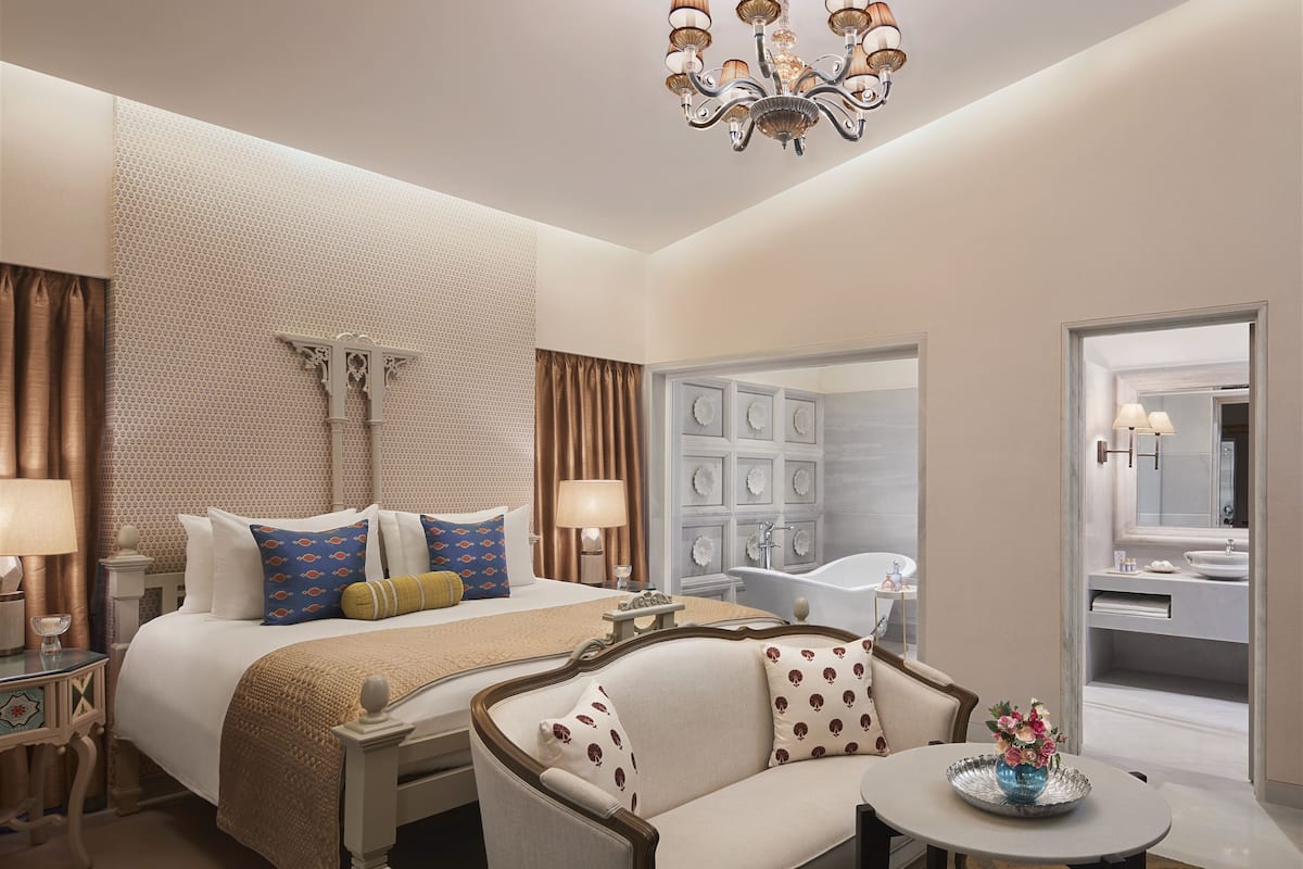 A hotel room with a large bed, a sofa, a round table, a chandelier, a bathroom with a bathtub, and a mirror.