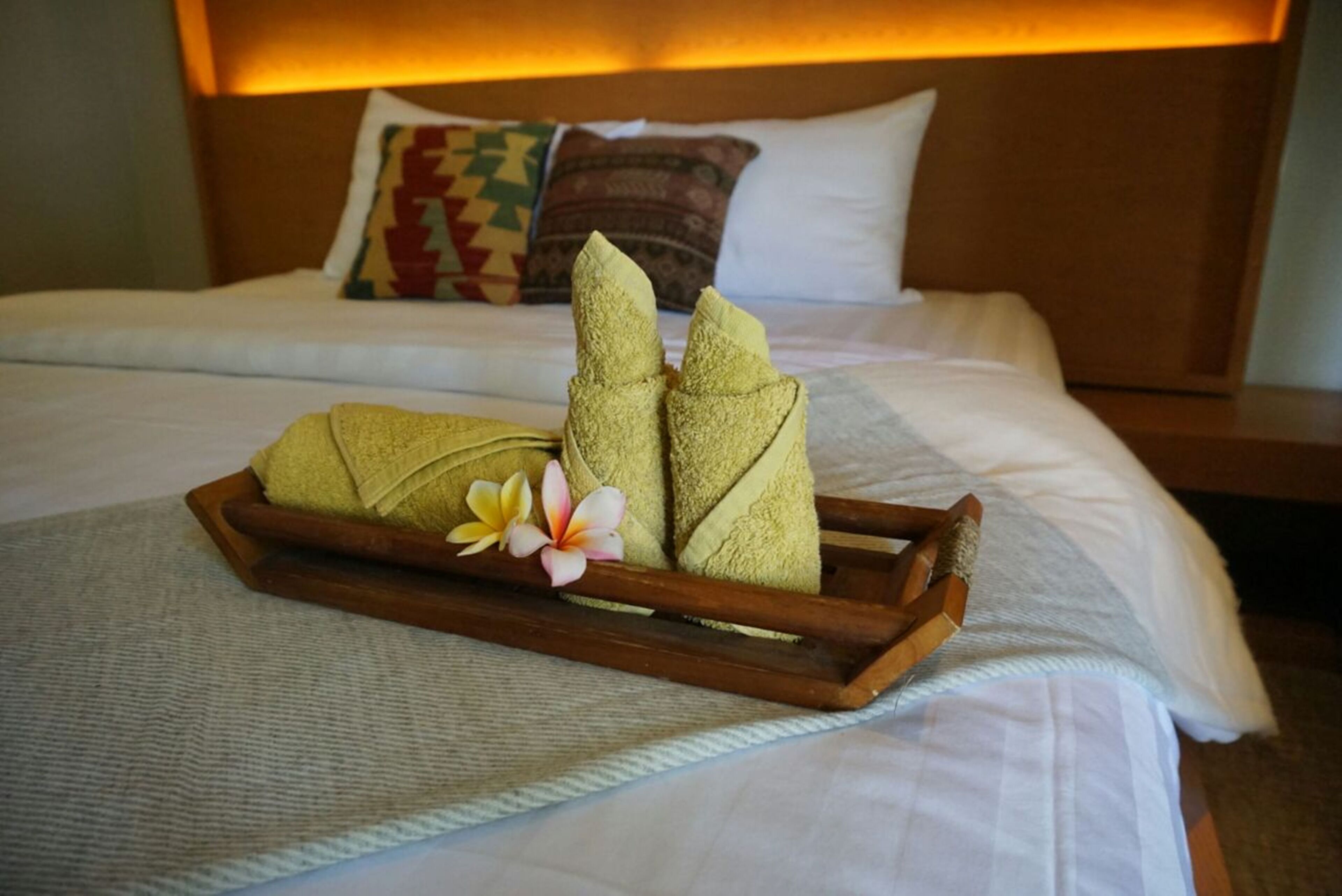 A neatly made bed with a wooden tray holding folded towels and a flower, set against a headboard with patterned pillows.