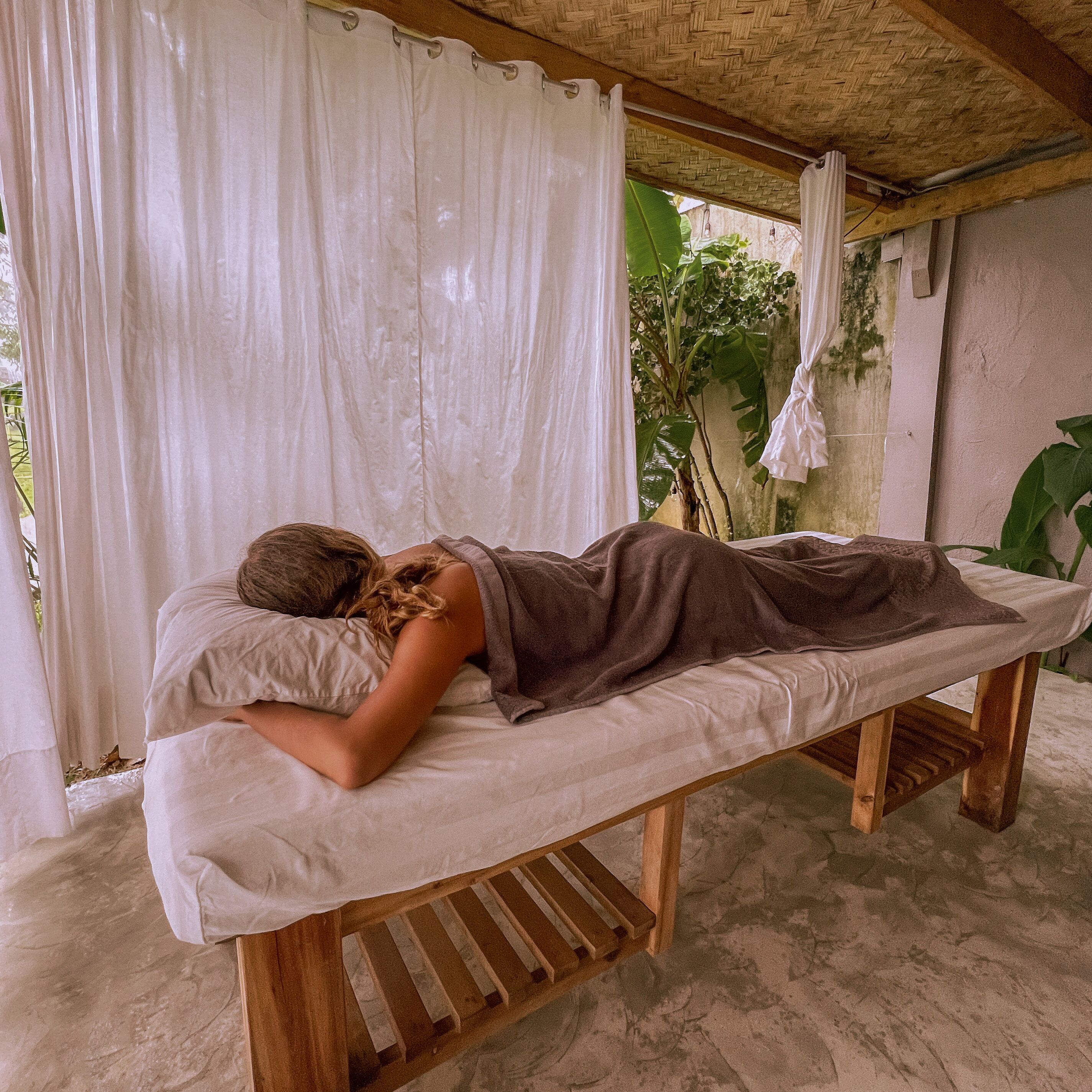 A person lying on a wooden bench under a thatched roof, with white curtains and greenery in the background.