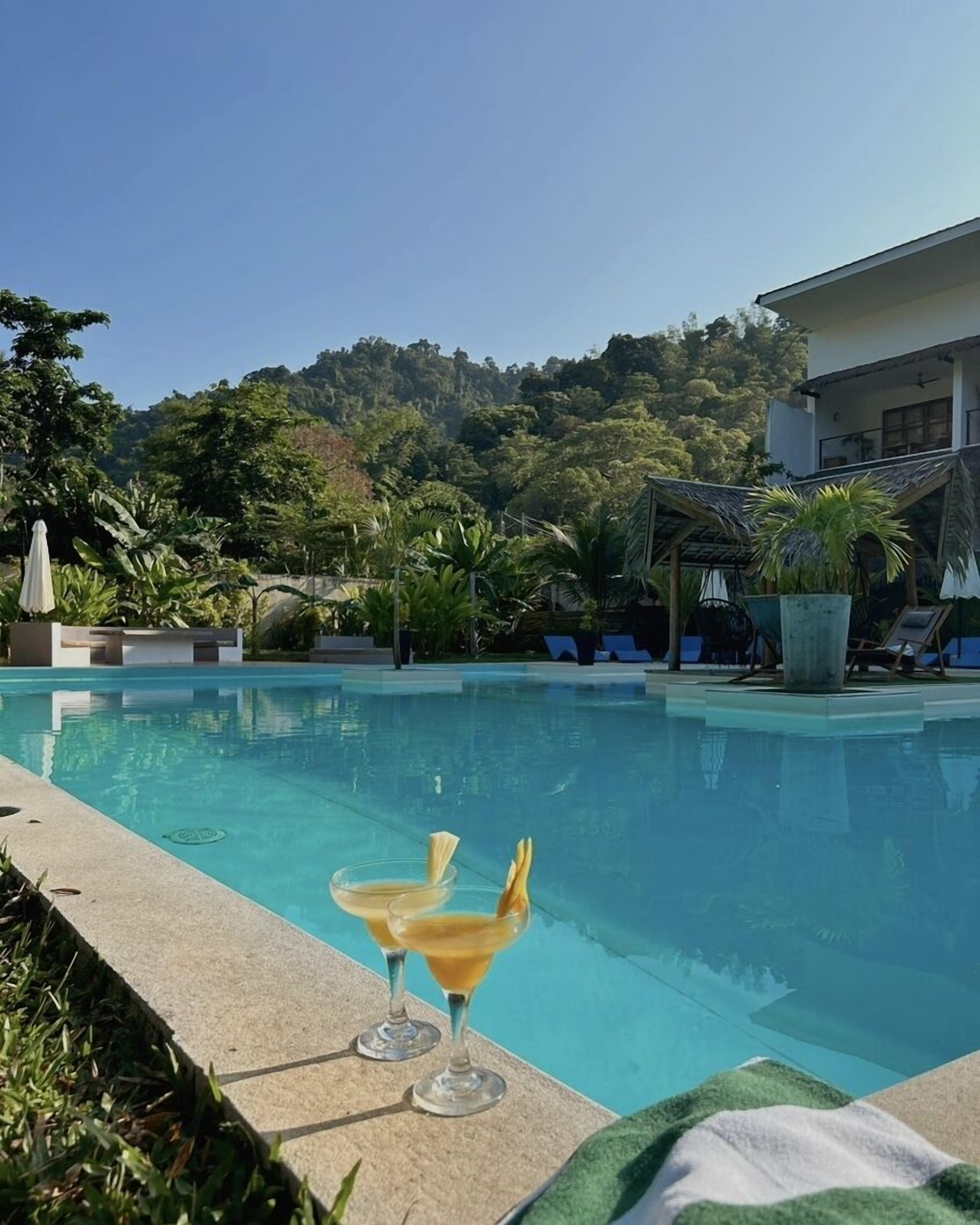 A swimming pool with two empty glasses of a yellow drink, surrounded by greenery and a building in the background.