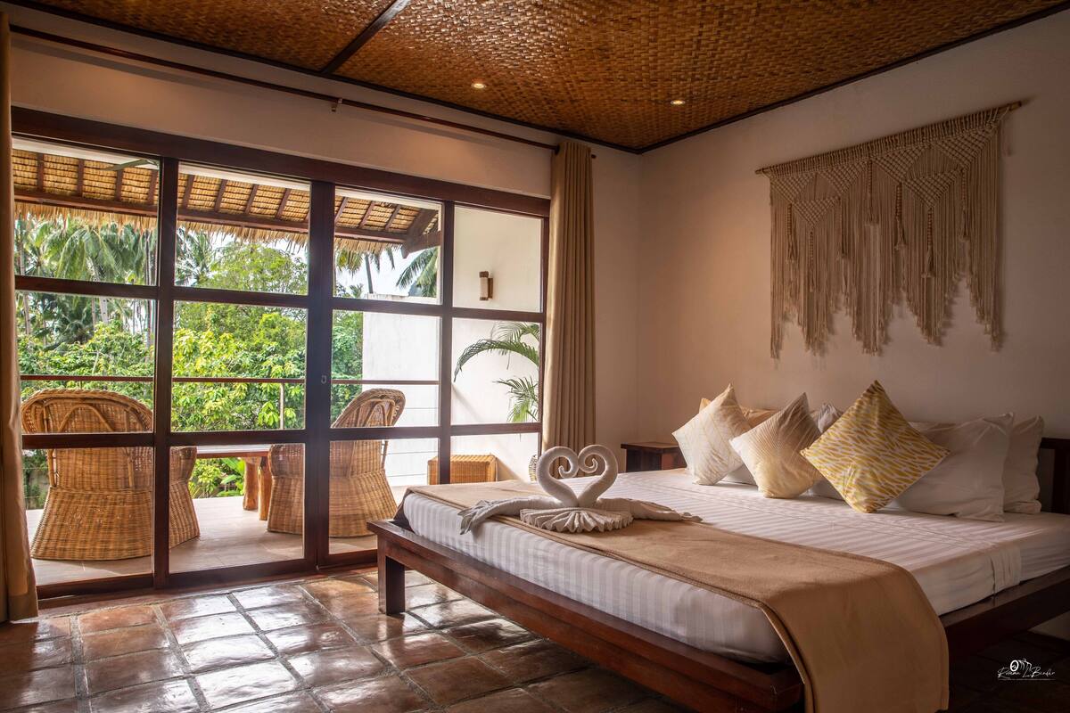 A bedroom with a large window, a bed with pillows, a bench, and a woven ceiling.