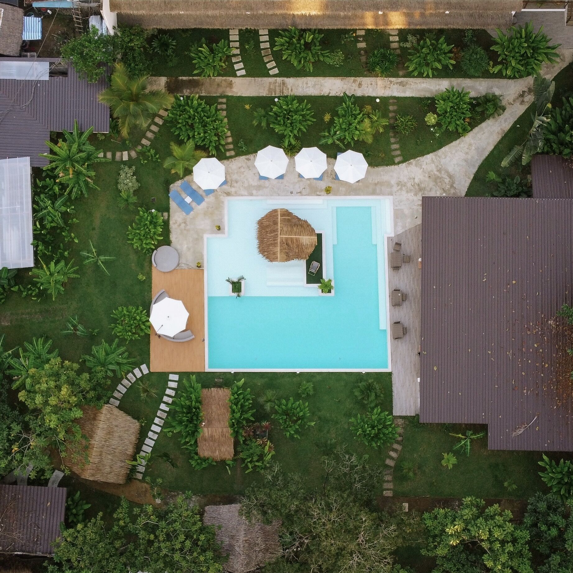 Vue aérienne d’un complexe hôtelier doté d’une piscine centrale, entourée de chaises longues et de parasols, ainsi que de diverses plantations.