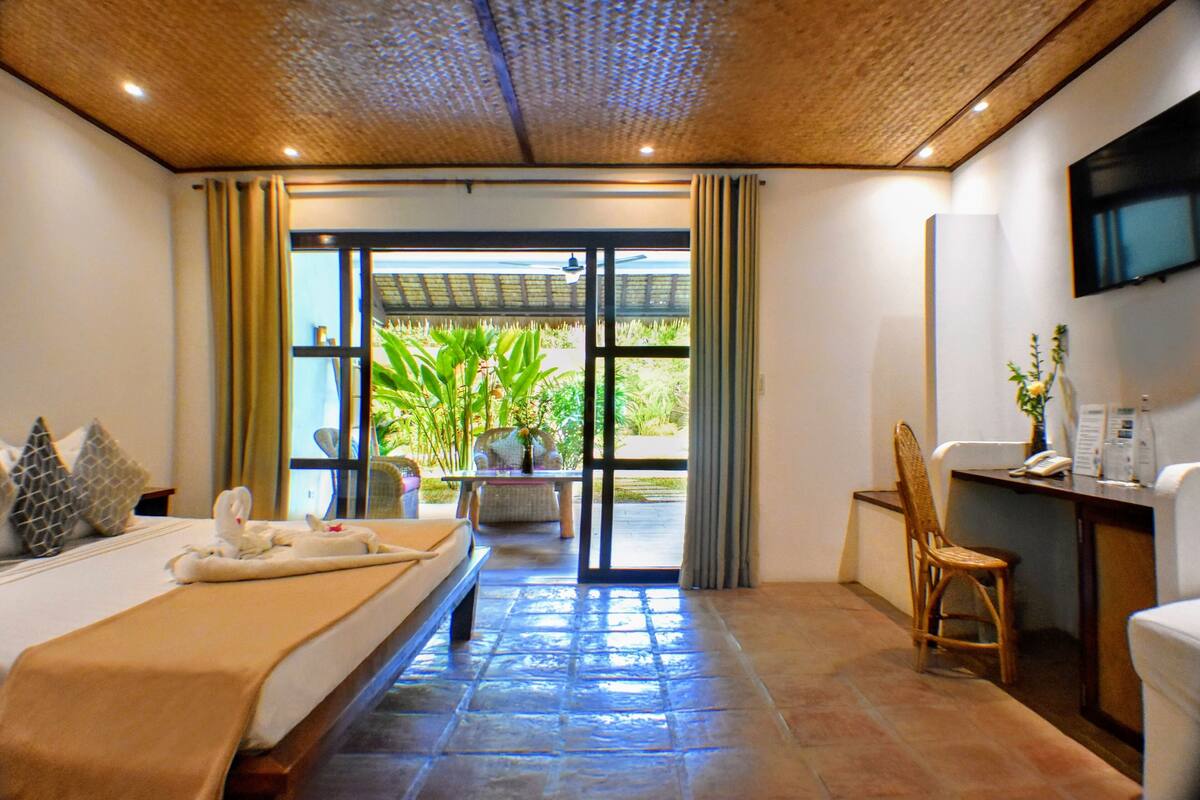 A hotel room with a bed, a desk, a chair, a TV, and a view of greenery outside.