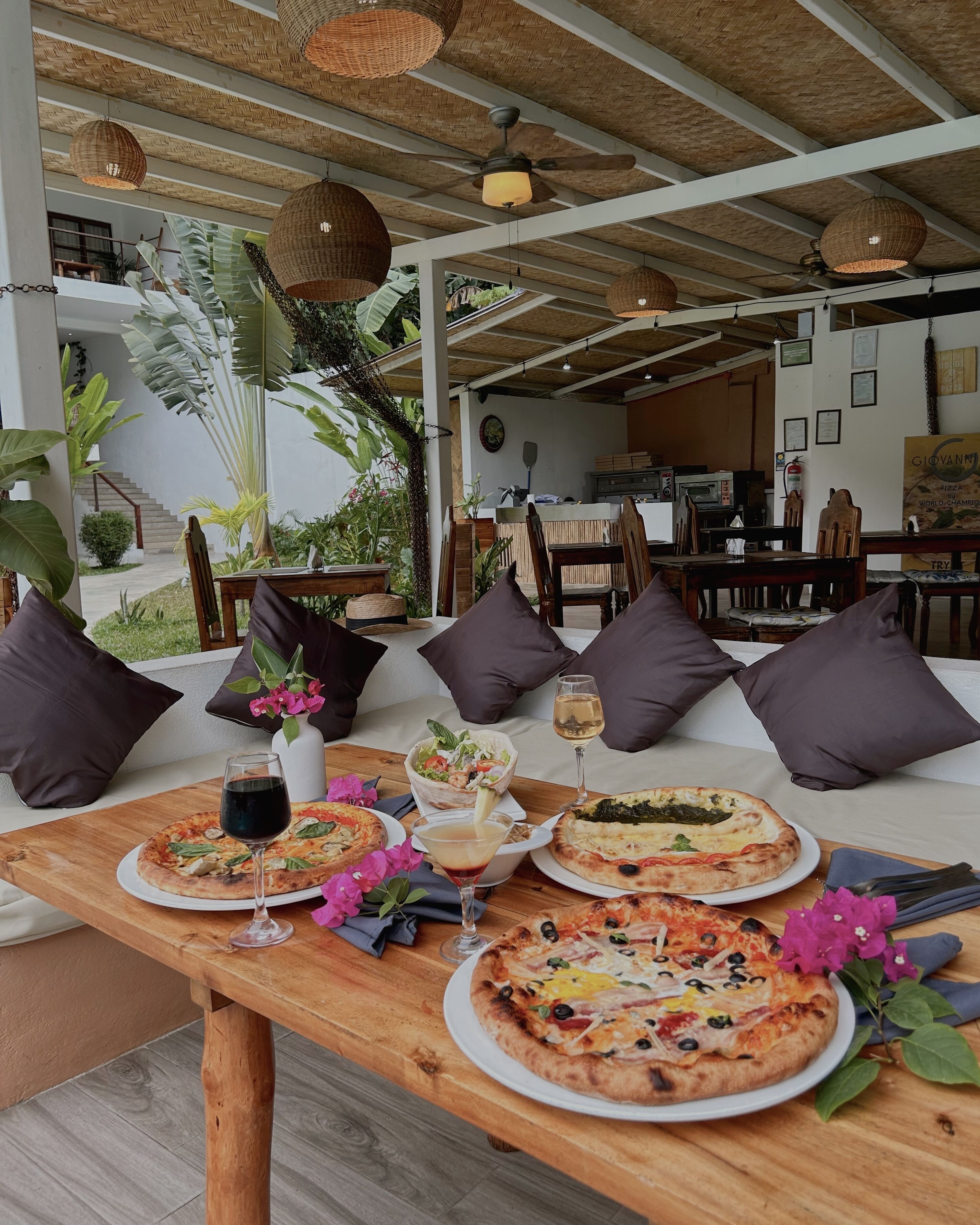 A dining area with a wooden table set with pizzas, drinks, and a salad. There are wicker hanging lights and a ceiling fan.