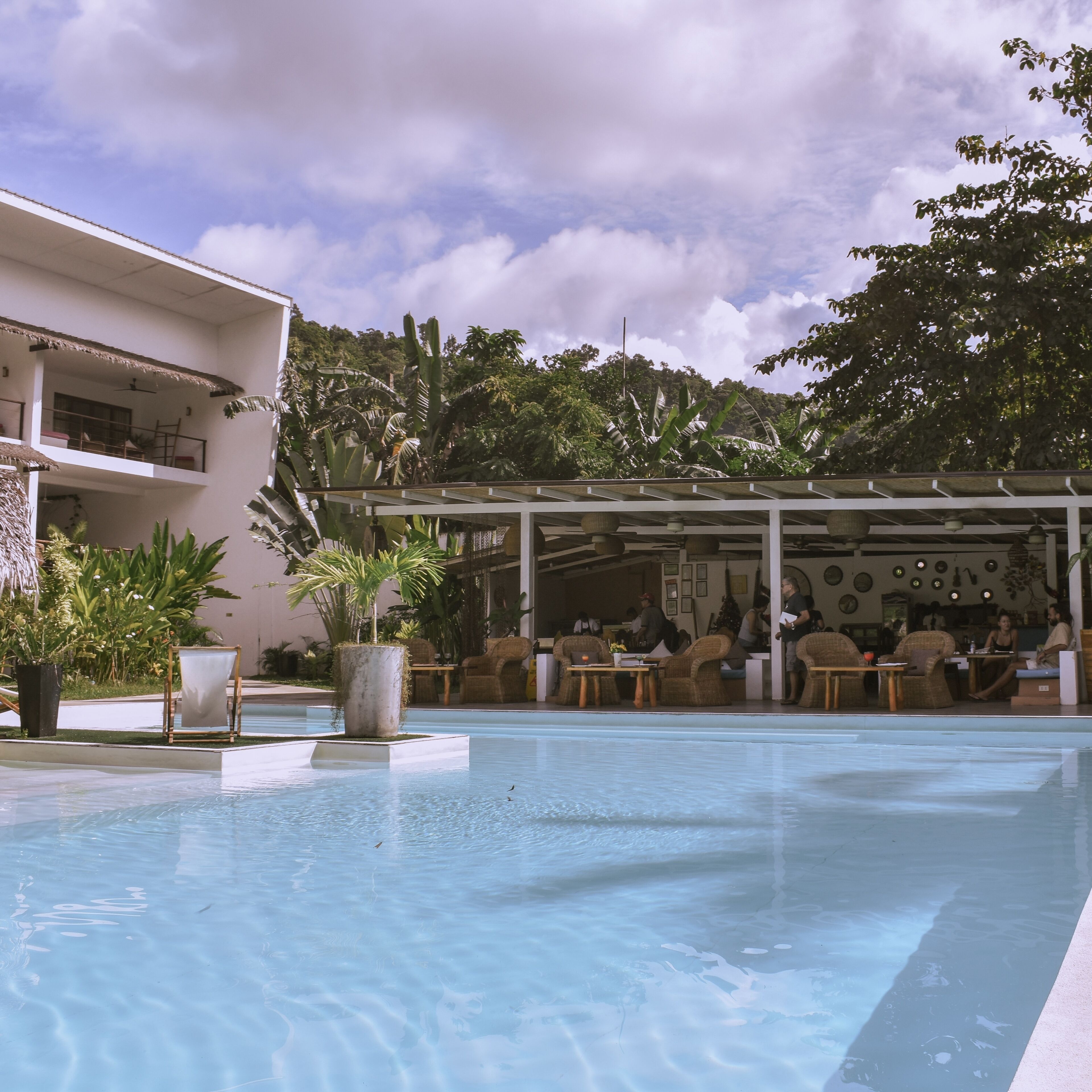 A swimming pool with lounge chairs and tables, surrounded by lush greenery and a building with a balcony.