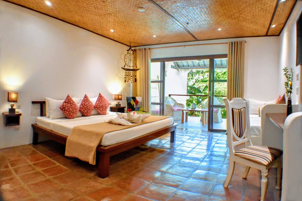 A hotel room with a large bed, a chair, a small table, and a balcony with a view of greenery.
