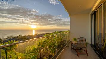 A balcony with a view of the sunset over the ocean, a glass railing, and two wicker chairs.