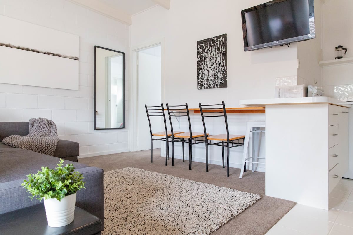 A modern living room with a sofa, a coffee table with a plant, a rug, and a dining area with chairs and a table.
