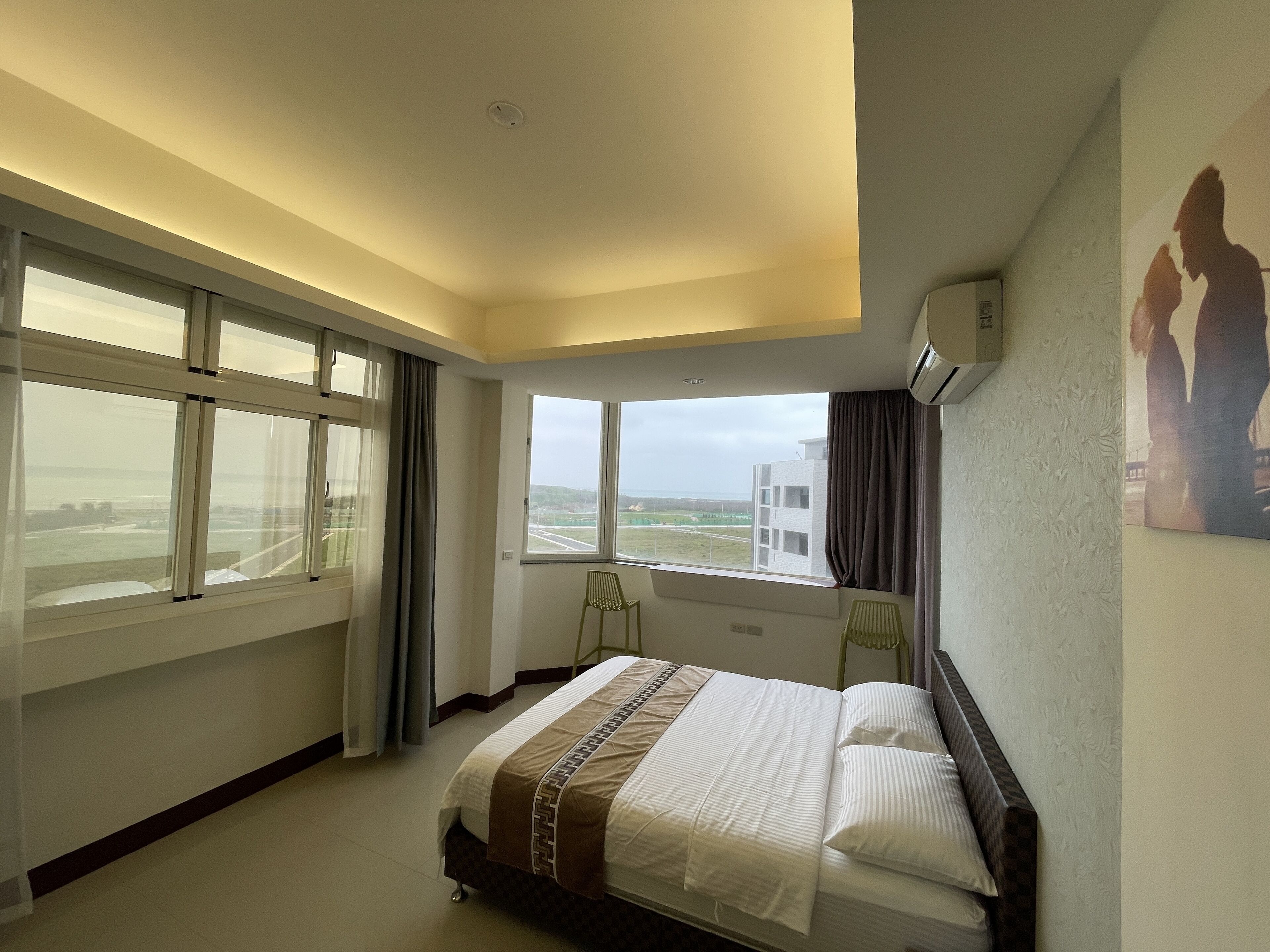 Una habitación de hotel con cama, una silla, un ventanal con vistas al aeropuerto y un equipo de aire acondicionado en la pared.
