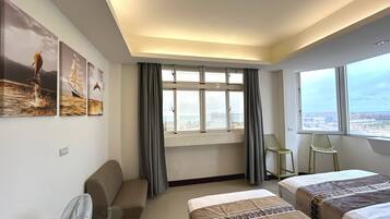 A hotel room with two beds, a desk, a chair, a window with a view, and artwork on the wall.