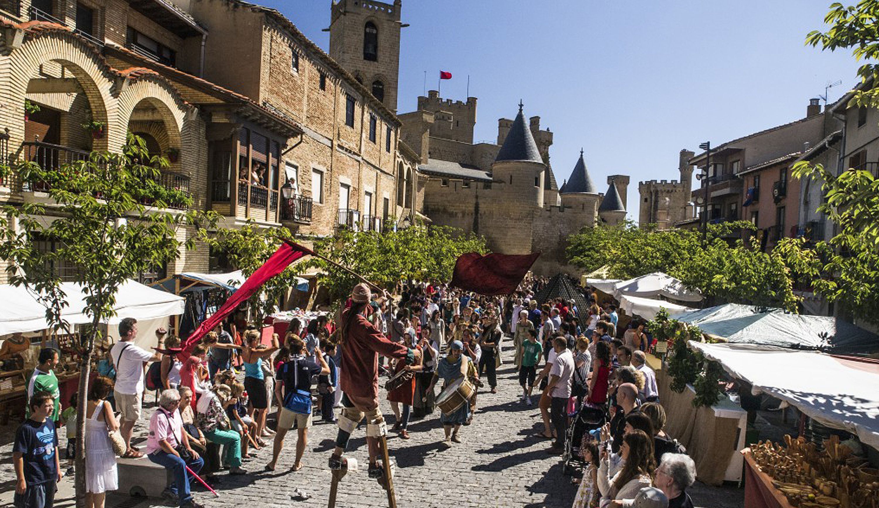 A bustling medieval street with stone buildings, a castle in the background, and people walking and gathering.
