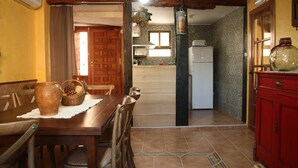 Un coin repas avec une table et des chaises en bois, un vase et un panier. On aperçoit un coin cuisine à l’arrière-plan, avec un réfrigérateur et une fenêtre.
