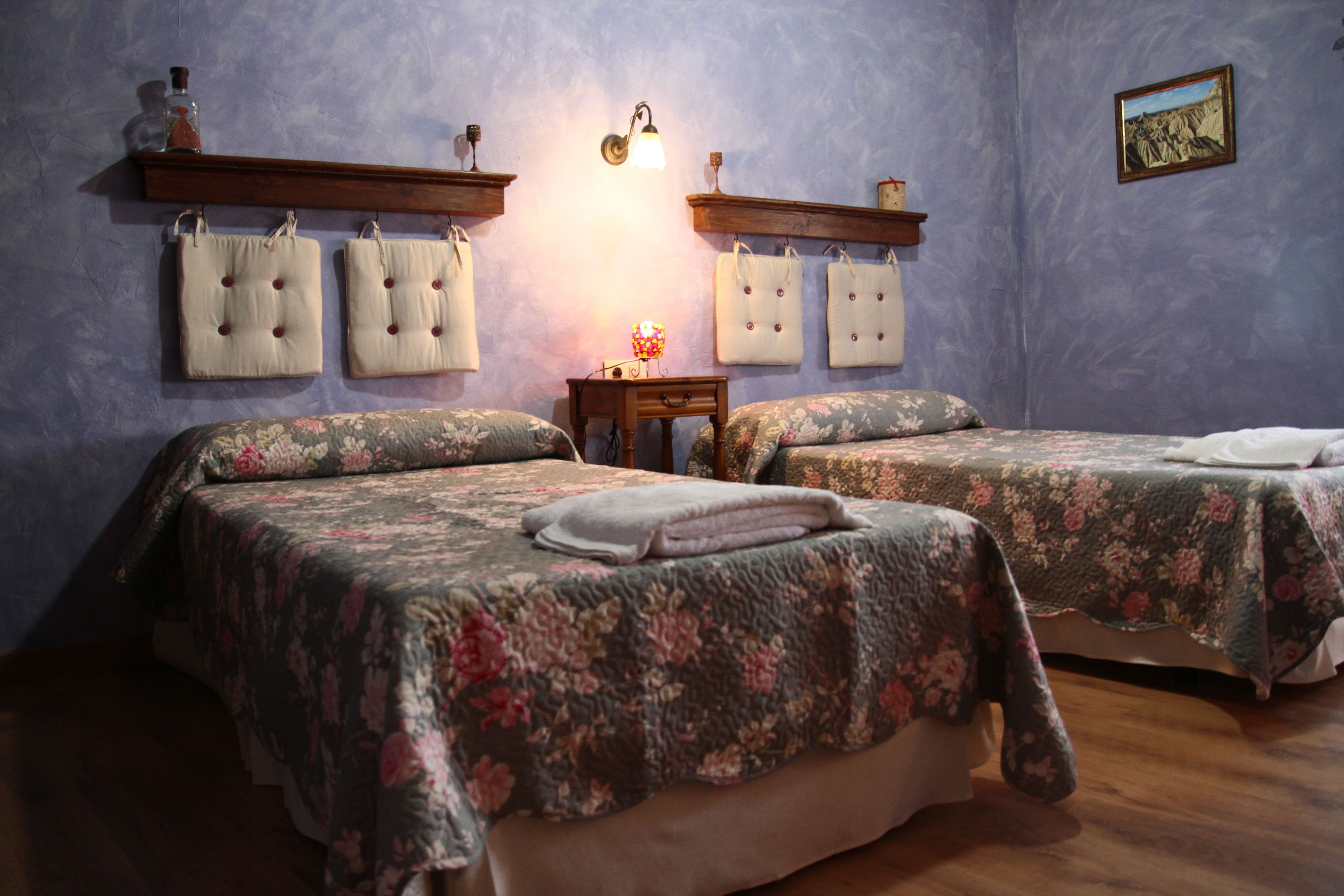 Ein Schlafzimmer mit zwei Betten, jeweils mit floralem Bettzeug und passenden Kissen. An der Wand sind Leuchten befestigt und es steht ein kleiner Tisch mit einer Lampe bereit.