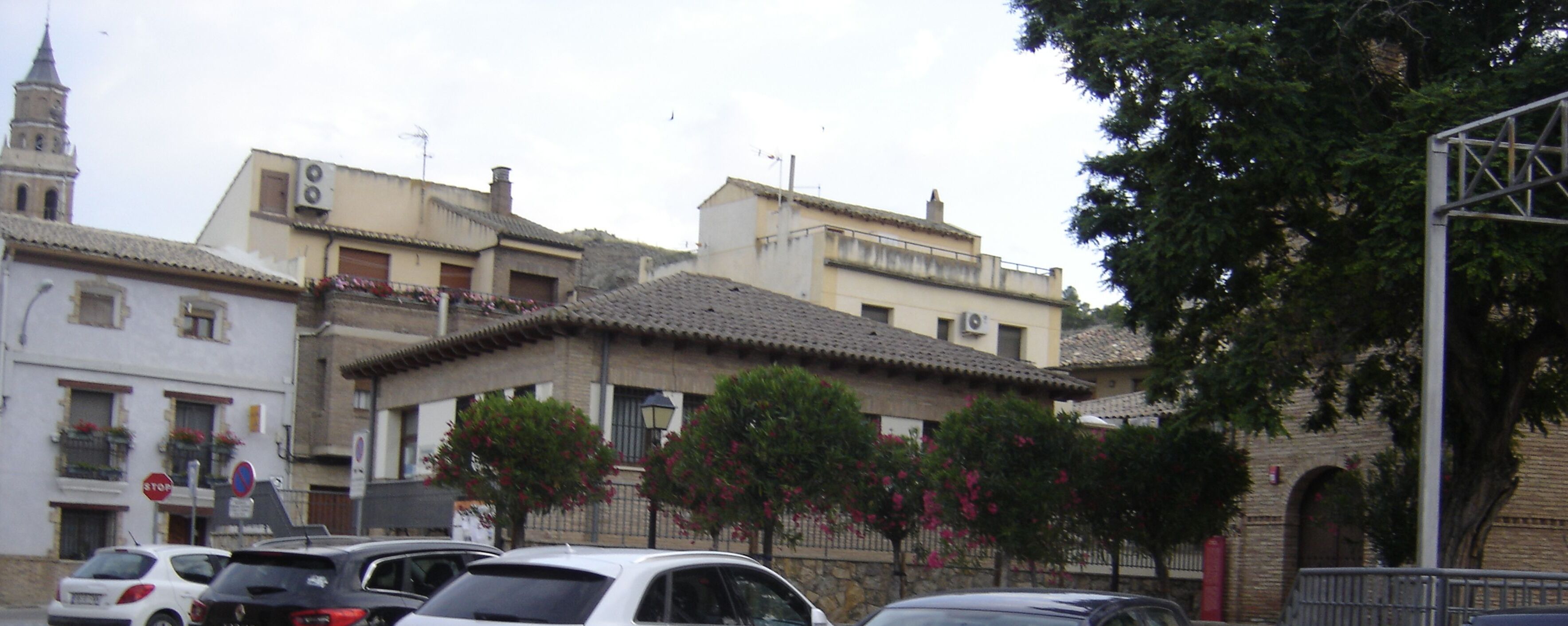 A street view with parked cars, buildings with tiled roofs, and a church tower.