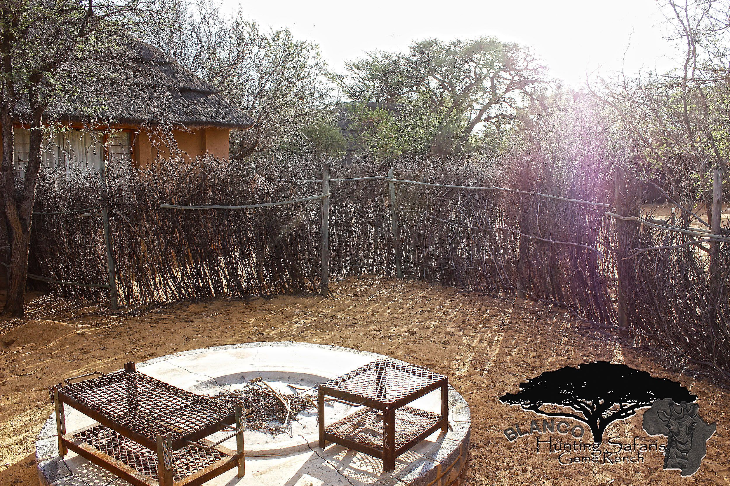 A fenced outdoor area with a thatched-roof structure, a fire pit, and a wooden table and chairs.