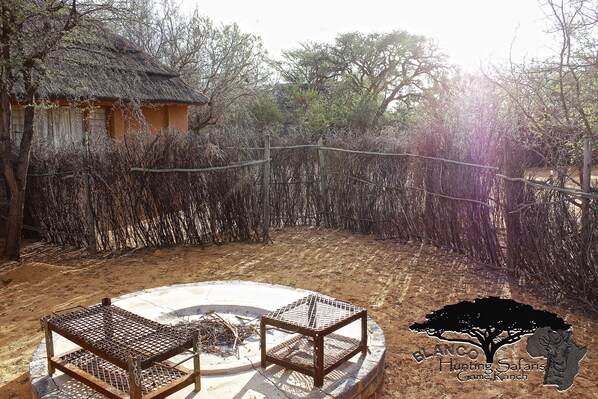 A fenced outdoor area with a thatched-roof structure, a fire pit, and a wooden table and chairs.