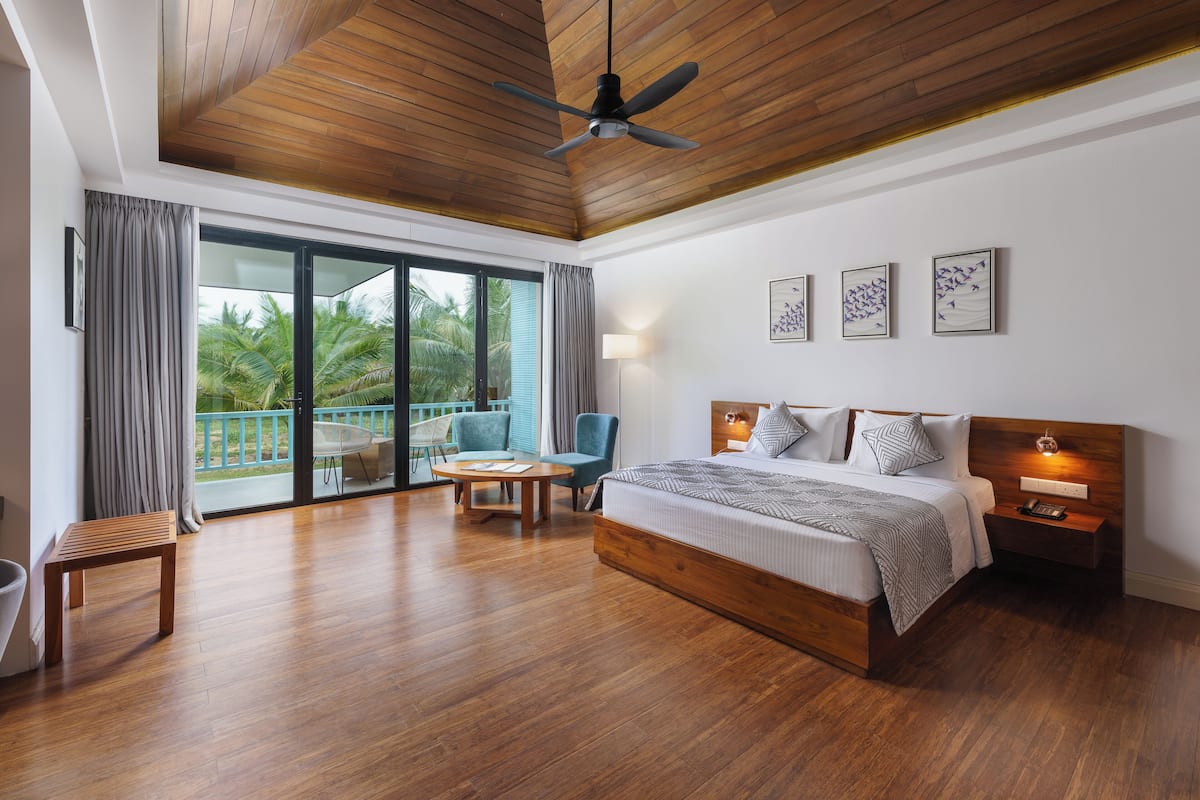 A modern bedroom with a large bed, a ceiling fan, a nightstand, and a view of palm trees through the glass door.