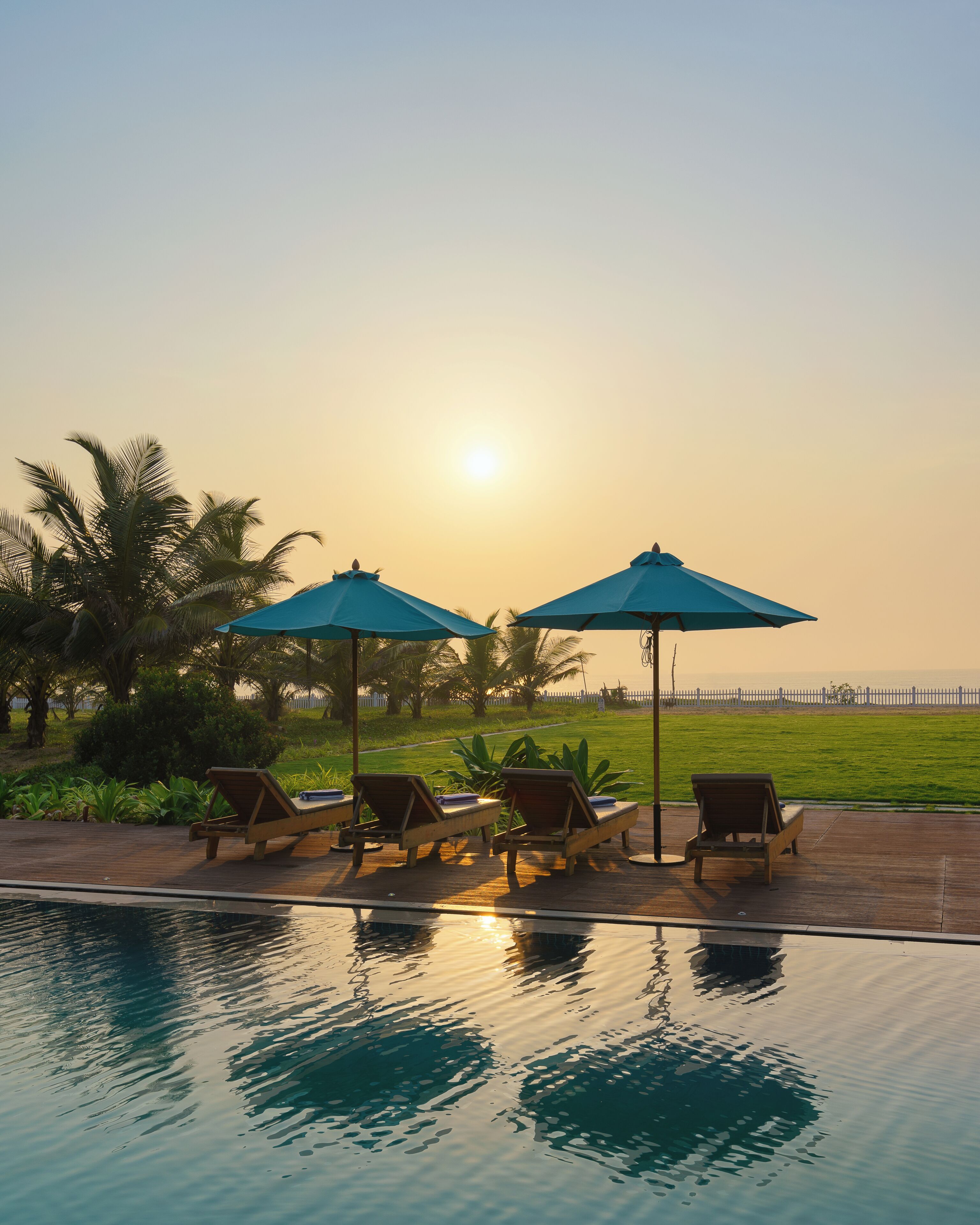 Zona junto a la piscina con sillones de descanso y sombrillas, vista despejada del atardecer y una piscina tranquila que refleja el cielo.
