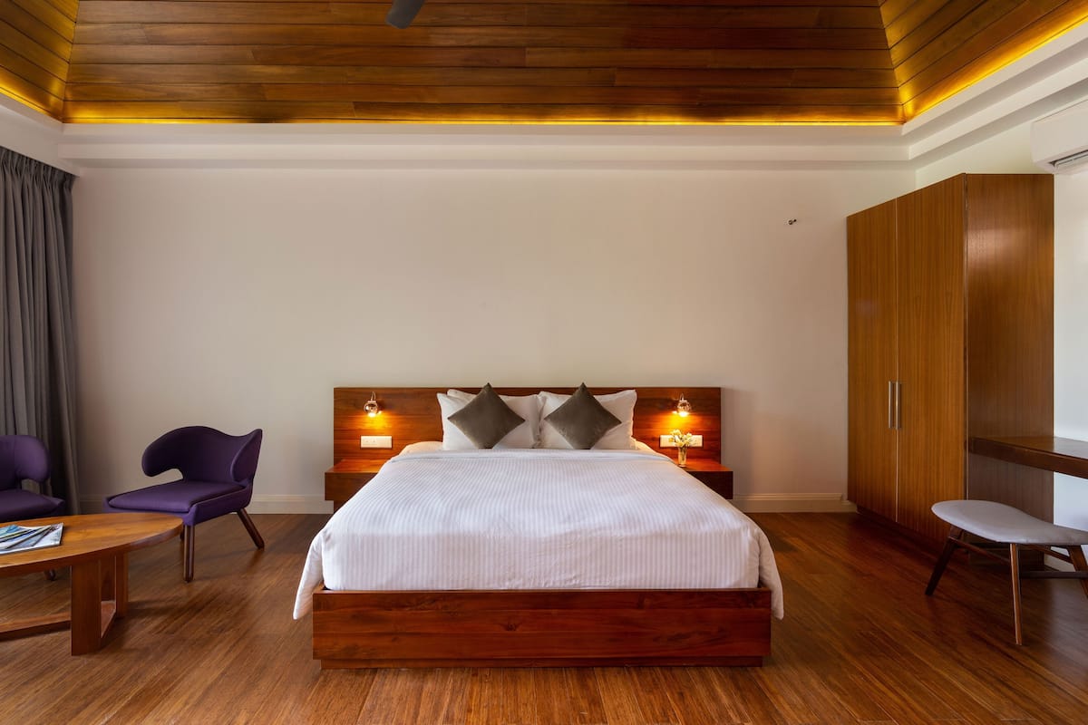 A modern hotel room with a wooden headboard, a large bed, a wooden wardrobe, and a desk with a chair.