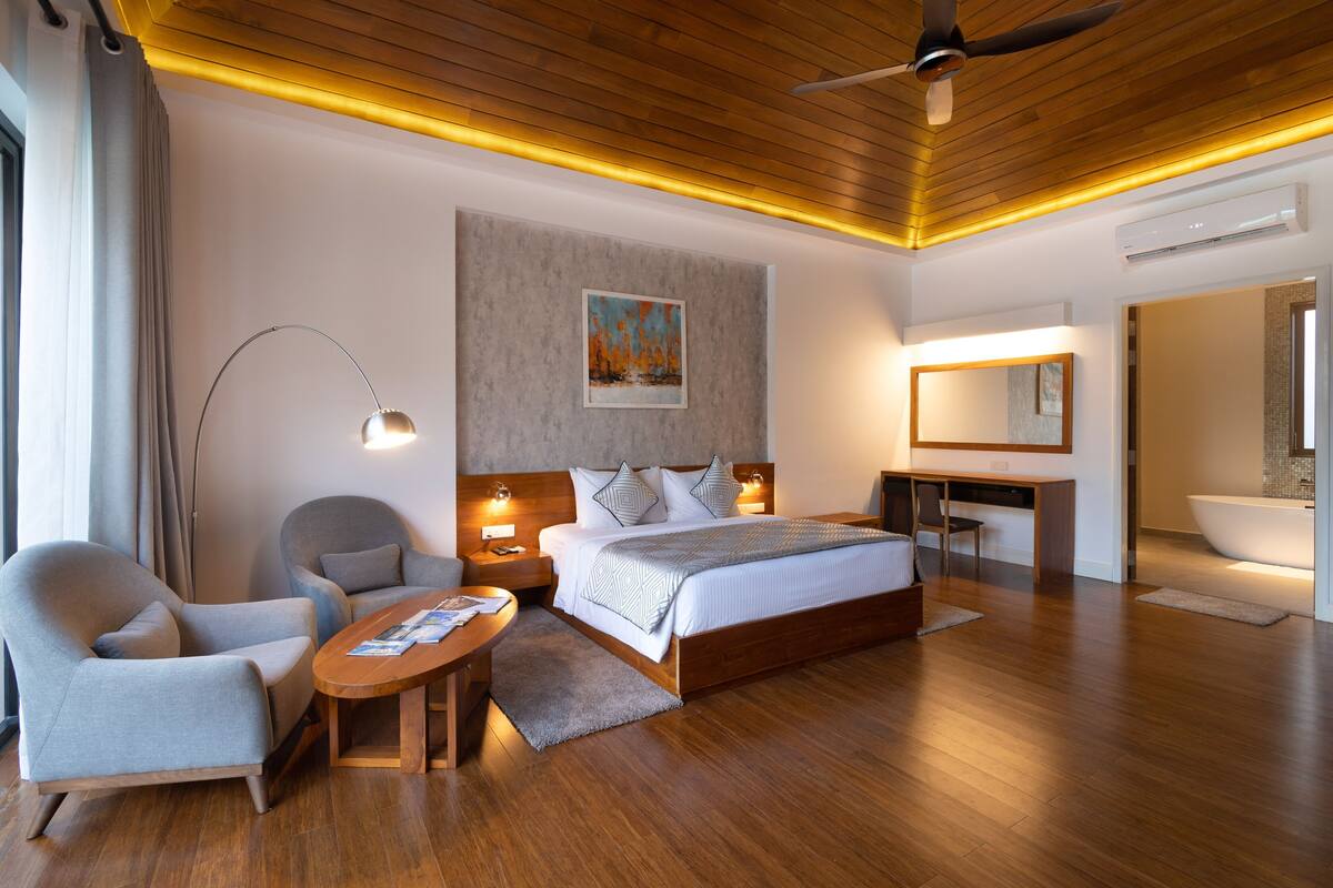 A modern hotel room with a wooden ceiling, a large bed, a desk, a chair, and a bathroom with a bathtub.