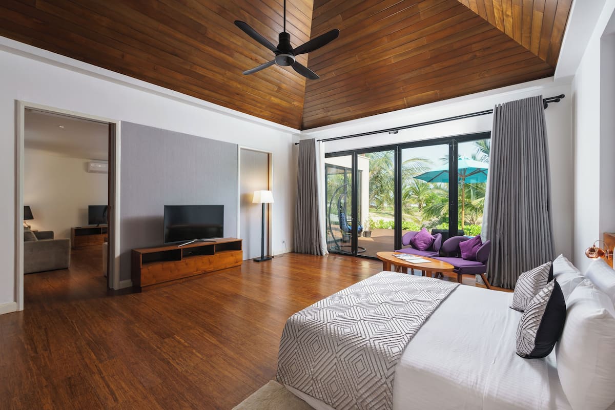 A spacious bedroom with a large bed, a TV, and a balcony with a view of palm trees.