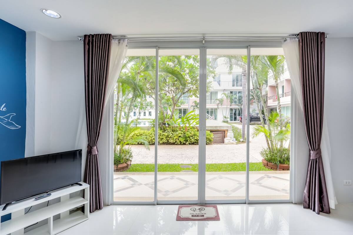 A modern living room with a flat-screen TV, a white entertainment unit, and a large sliding glass door leading to a courtyard with greenery and a building in the background.