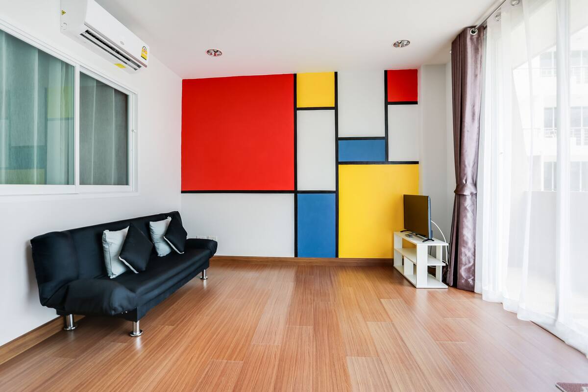 A modern living room with a black sofa, a colorful geometric wall, and a wooden floor.