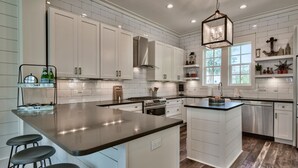 A modern kitchen with white cabinetry, a central island with bar stools, stainless steel appliances, and a hanging light fixture. - New construction, custom decorated, chic and spacious Seacrest home, sleeps 14 (Rosemary Beach)