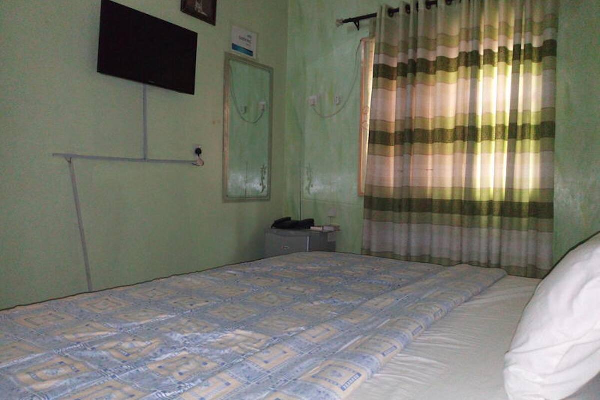A room with a bed, a television mounted on the wall, a window with curtains, and a small table with a lamp.