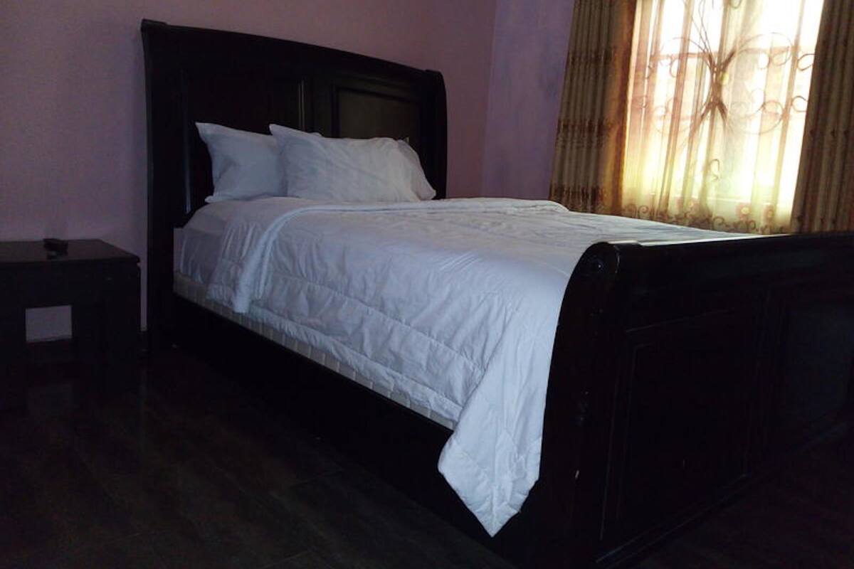 A single bed with a dark headboard in a room with a plain wall and a window with curtains.