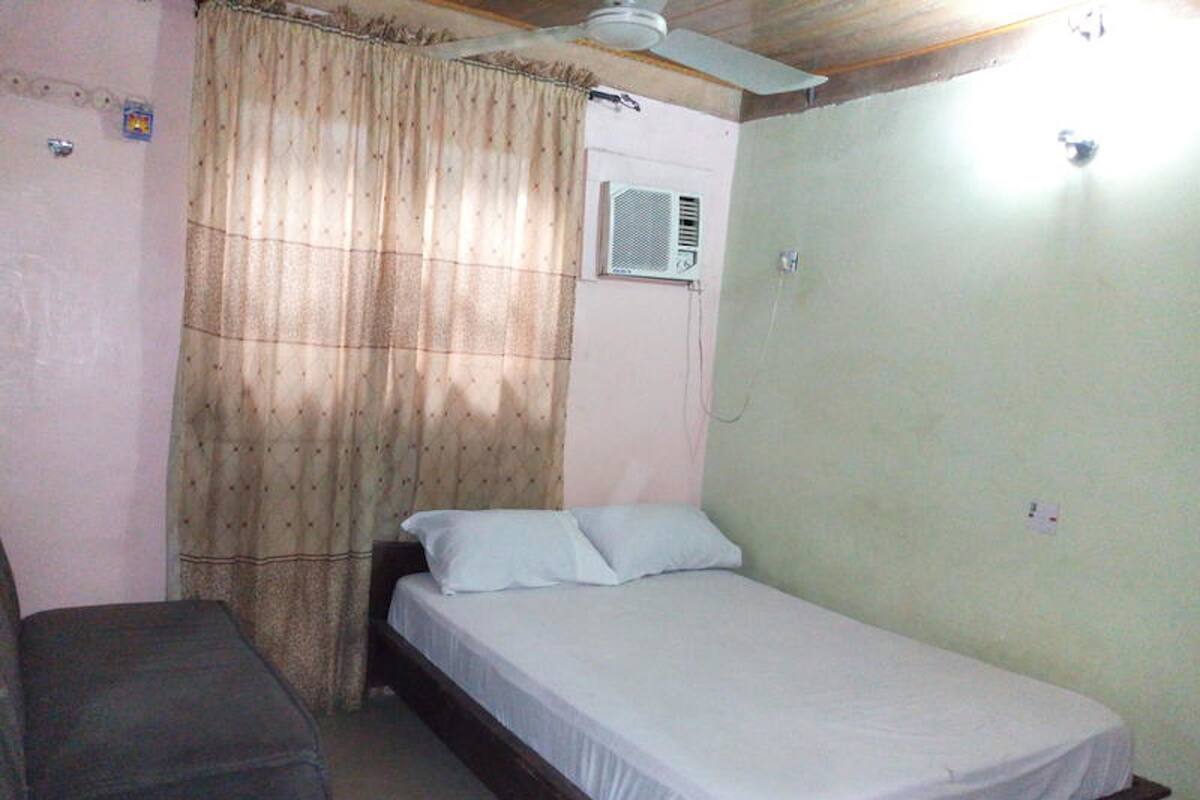 A small, single-bed room with a ceiling fan, a window with curtains, and an air conditioning unit.
