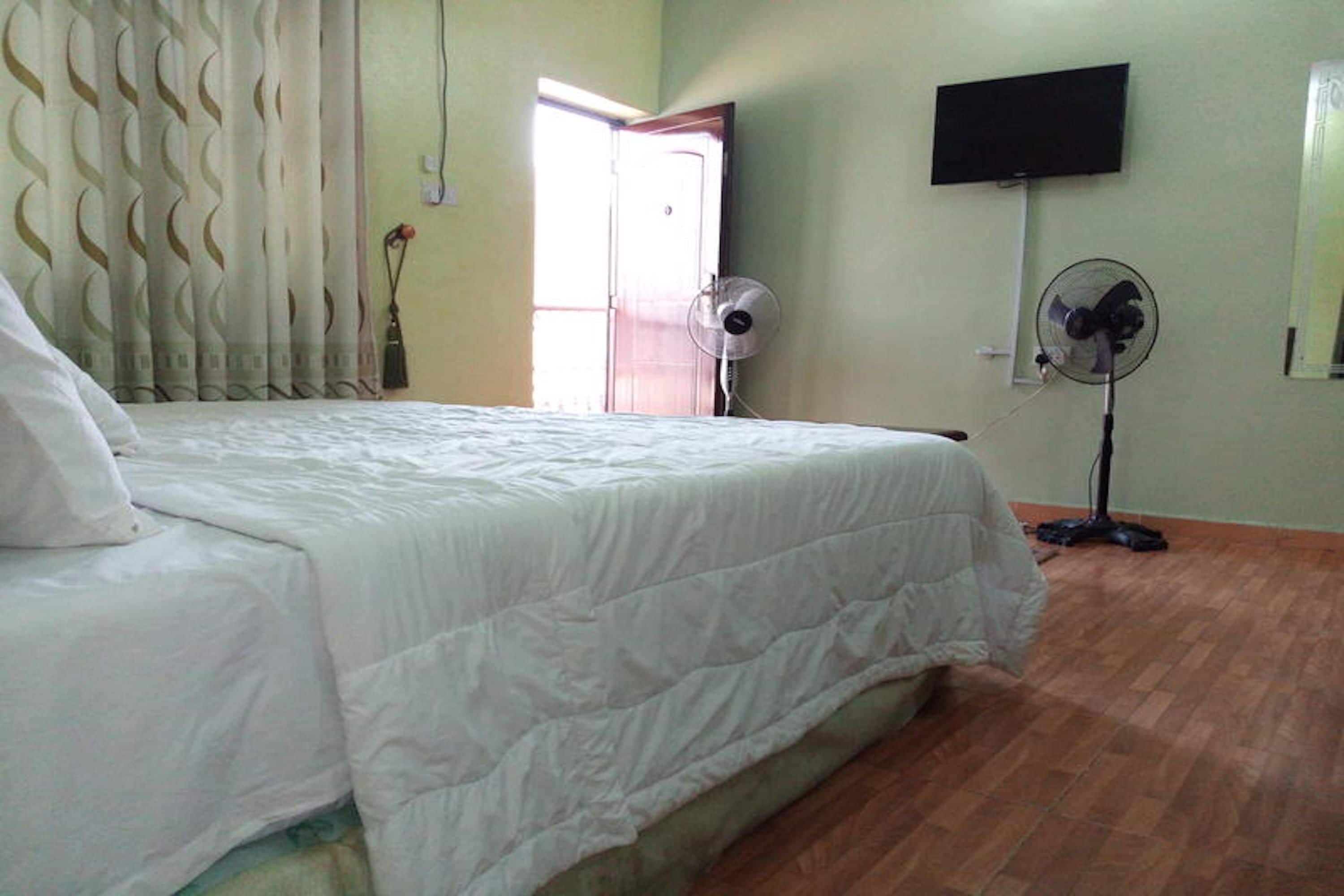 A bedroom with a bed, a standing fan, a television on the wall, and a door leading to another room.