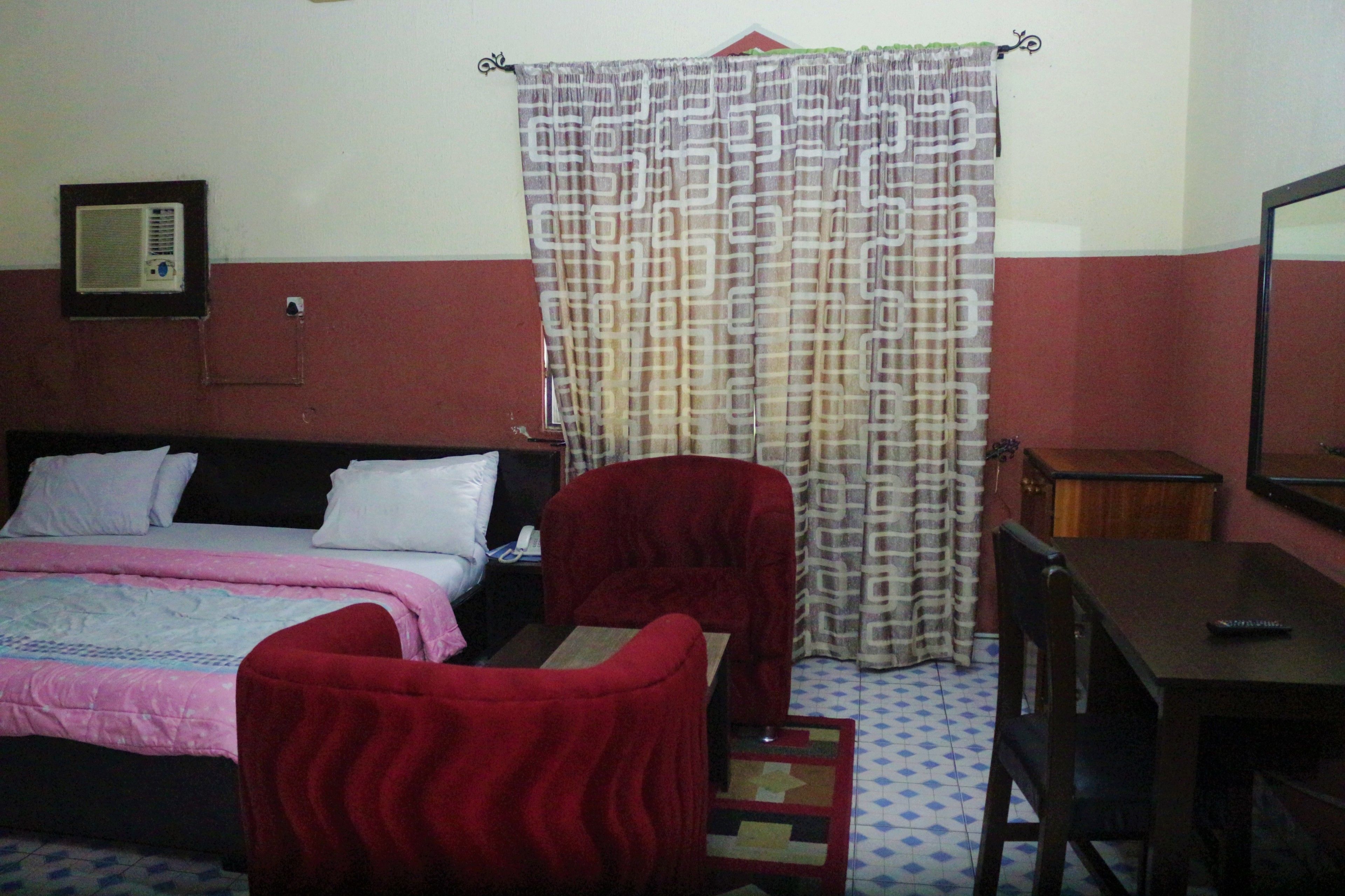 A bedroom with a bed, two red chairs, a wooden table, and a curtain.