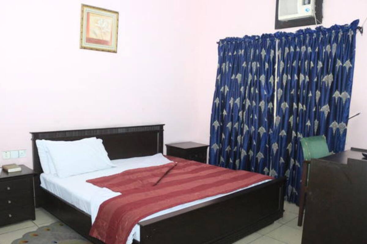 A bedroom with a bed, a nightstand, a bedside table, a chair, a desk, a window with curtains, and a wall-mounted air conditioner.