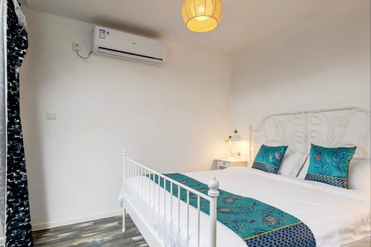 Une chambre avec un lit blanc, une couvre-lit à motifs turquoise et bleu, une tête de lit blanche, une table de chevet avec une lampe et un climatiseur fixé au mur.
