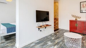 Une chambre d’hôtel moderne avec un téléviseur à écran plat fixé au mur, un meuble rouge agrémenté d’objets décoratifs, une petite table d’appoint ronde et blanche, et un parquet à motifs.