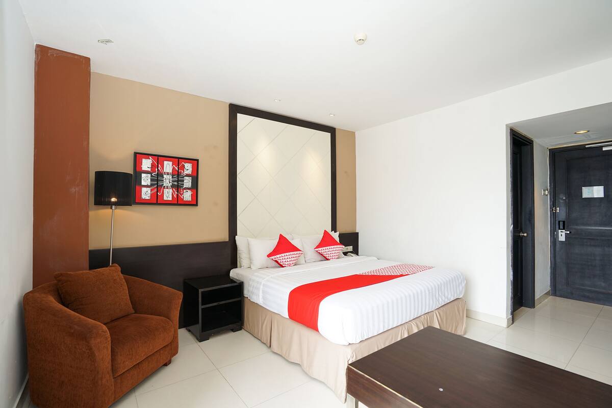 Une chambre d’hôtel avec un lit, un fauteuil brun, une table de chevet et un grand miroir au mur.