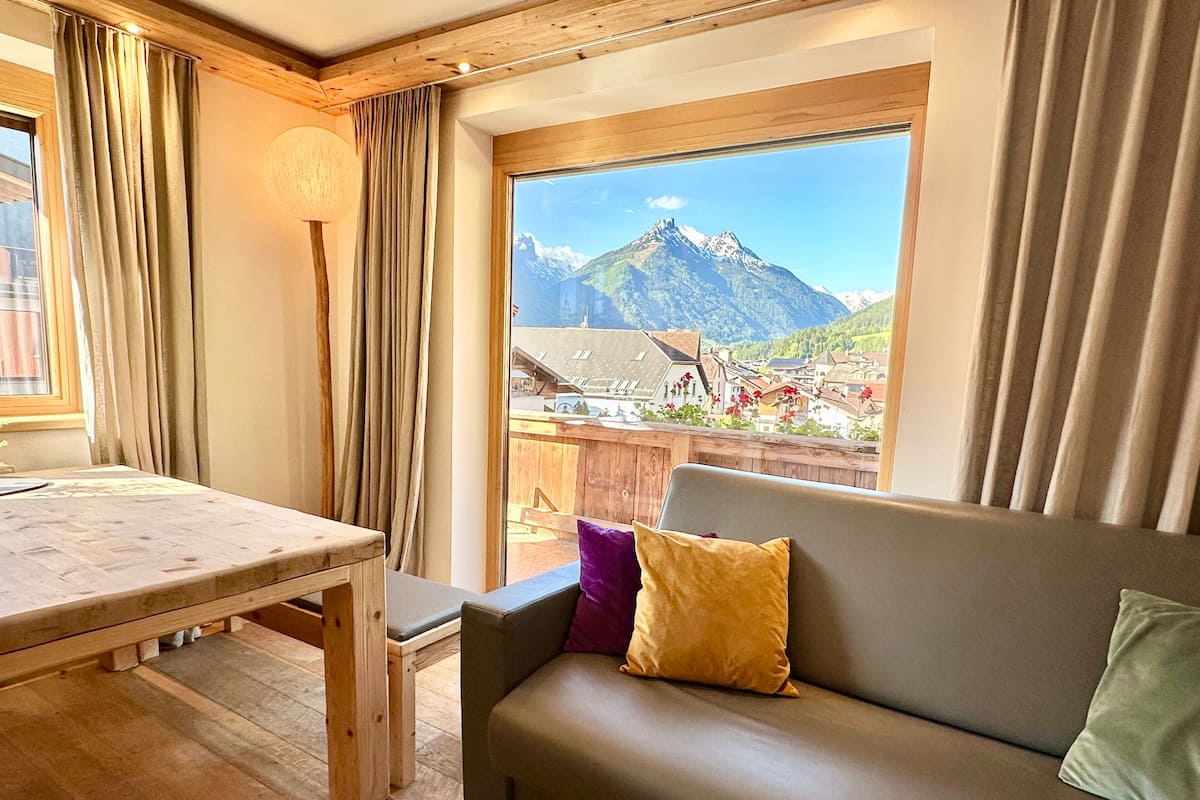 A room with a wooden table, a sofa with cushions, and a large window offering a mountain view.